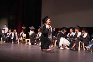 A young person in a graduation gown and cap dances joyfully on stage while peers in similar attire sit nearby.