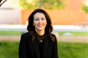 A smiling person with dark hair and glasses wears a black blazer and necklace, standing outdoors in front of green grass and a blurred orange wall.