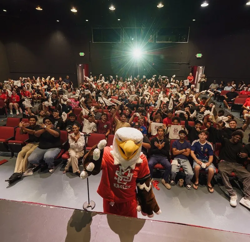 An eagle mascot stands on a stage in front of a large audience seated in an auditorium, many holding up white items.