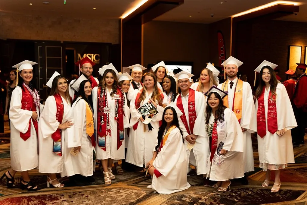 A group of graduates in white gowns and caps pose together, wearing red sashes and accessories, at a celebratory event.